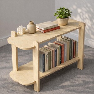 May include: A light-coloured wooden bookshelf with two shelves. The top shelf holds books, a small potted plant, a candle, and a vase. The bottom shelf is filled with books. The bookshelf has a simple, modern design.