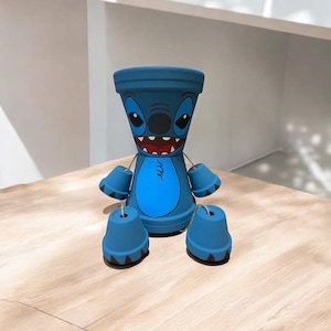 Può includere: Vaso da fiori in ceramica blu a forma di Stitch di Lilo & Stitch. Il vaso ha quattro piccoli vasi attaccati ai lati, creando un design fantasioso.