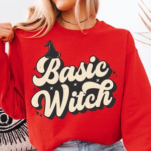 May include: A red sweatshirt with the words "Basic Witch" in a retro font, with a witch hat graphic. The sweatshirt is a solid red color and has long sleeves. The text is in black and cream colors.