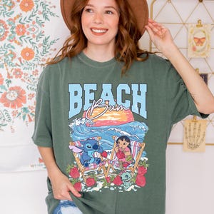 A smiling woman wearing a green t-shirt with a beach scene and cartoon characters, standing in front of a floral wall.