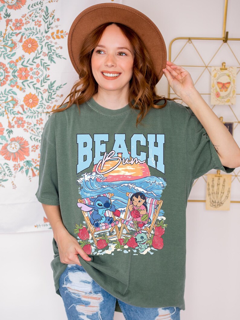 A smiling woman wearing a green t-shirt with a beach scene and cartoon characters, standing in front of a floral wall.