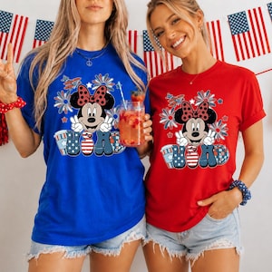 May include: Two women wearing blue and red t-shirts with a Minnie Mouse graphic. The graphic features Minnie Mouse with a bow, peace signs, and the letters "USA" in patriotic colors. American flags and decorations are in the background.