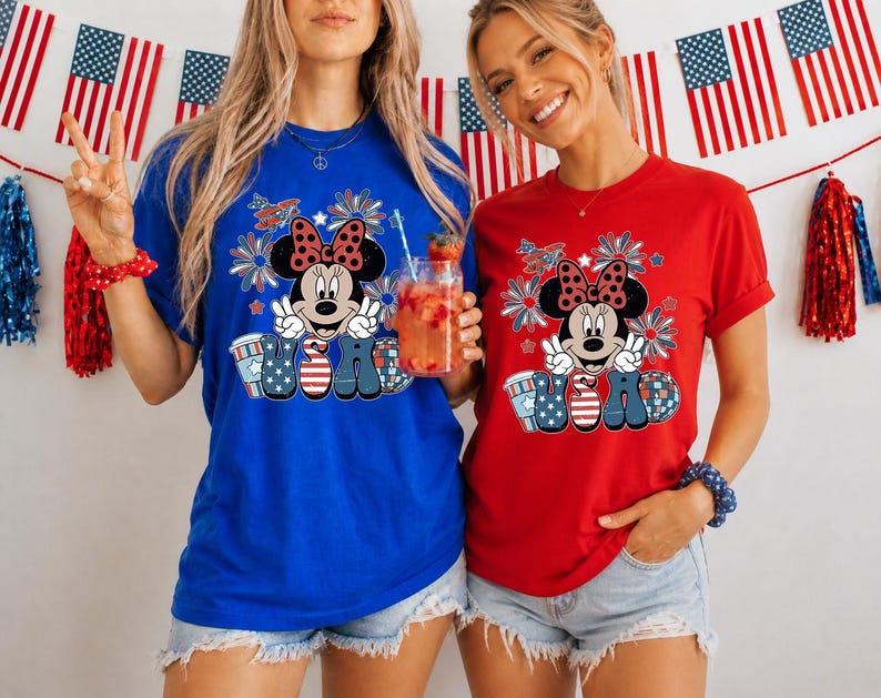 May include: Two women wearing blue and red t-shirts with a Minnie Mouse graphic. The graphic features Minnie Mouse with a bow, peace signs, and the letters "USA" in patriotic colors. American flags and decorations are in the background.