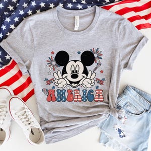 May include: A gray t-shirt featuring a graphic of Mickey Mouse giving a peace sign, with fireworks and the word "AMERICA" in red, white, and blue. The shirt is paired with denim shorts and white sneakers, with an American flag in the background.