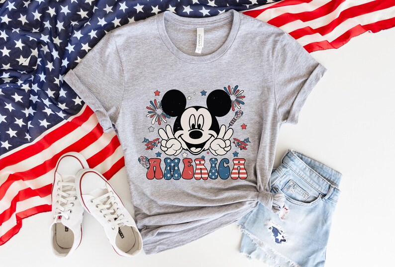 May include: A gray t-shirt featuring a graphic of Mickey Mouse giving a peace sign, with fireworks and the word "AMERICA" in red, white, and blue. The shirt is paired with denim shorts and white sneakers, with an American flag in the background.