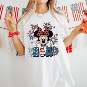 May include: White t-shirt featuring a Minnie Mouse graphic with a red bow, fireworks, and the word "USA" in patriotic colors. The design includes a coffee cup, a disco ball, and an airplane. American flags and decorations are in the background.
