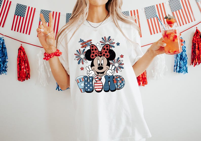 May include: White t-shirt featuring a Minnie Mouse graphic with a red bow, fireworks, and the word "USA" in patriotic colors. The design includes a coffee cup, a disco ball, and an airplane. American flags and decorations are in the background.