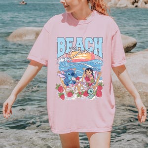 A woman wearing a pink t-shirt with a graphic design of a cartoon character and the text "BEACH Bum" printed on it, standing near the water.