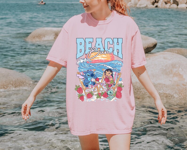 A woman wearing a pink t-shirt with a graphic design of a cartoon character and the text "BEACH Bum" printed on it, standing near the water.