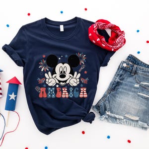 May include: Navy blue t-shirt featuring Mickey Mouse with peace signs and the word "AMERICA" in red, white, and blue. The image includes denim shorts, a red bandana, and decorative fireworks, perfect for Independence Day celebrations.