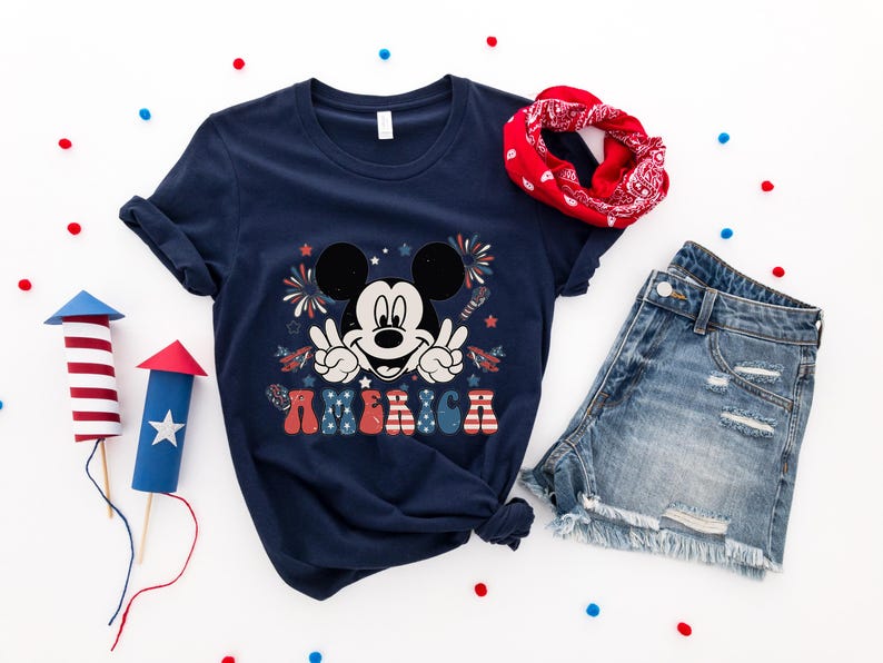 May include: Navy blue t-shirt featuring Mickey Mouse with peace signs and the word "AMERICA" in red, white, and blue. The image includes denim shorts, a red bandana, and decorative fireworks, perfect for Independence Day celebrations.