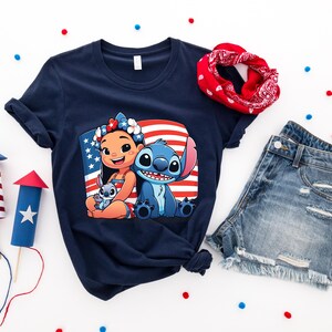 May include: Navy blue t-shirt featuring a graphic of Lilo and Stitch with an American flag background. The image is surrounded by a red bandana, denim shorts, and decorative fireworks. Red, white, and blue confetti dots the white background.