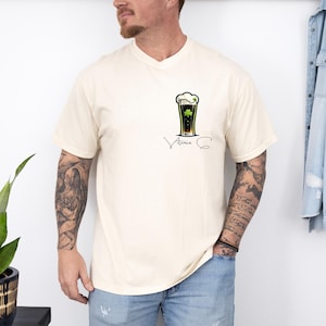 A man wearing a white t-shirt with a graphic of a beer glass and a clover, standing next to a plant and a denim jacket.