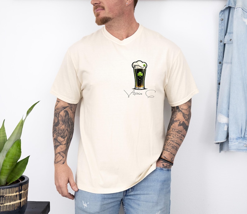 A man wearing a white t-shirt with a graphic of a beer glass and a clover, standing next to a plant and a denim jacket.