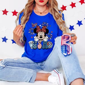 May include: A royal blue t-shirt featuring a Minnie Mouse graphic with a red bow, peace signs, and the letters "USA" in patriotic colors. The person is wearing light blue jeans and holding red, white, and blue sunglasses.