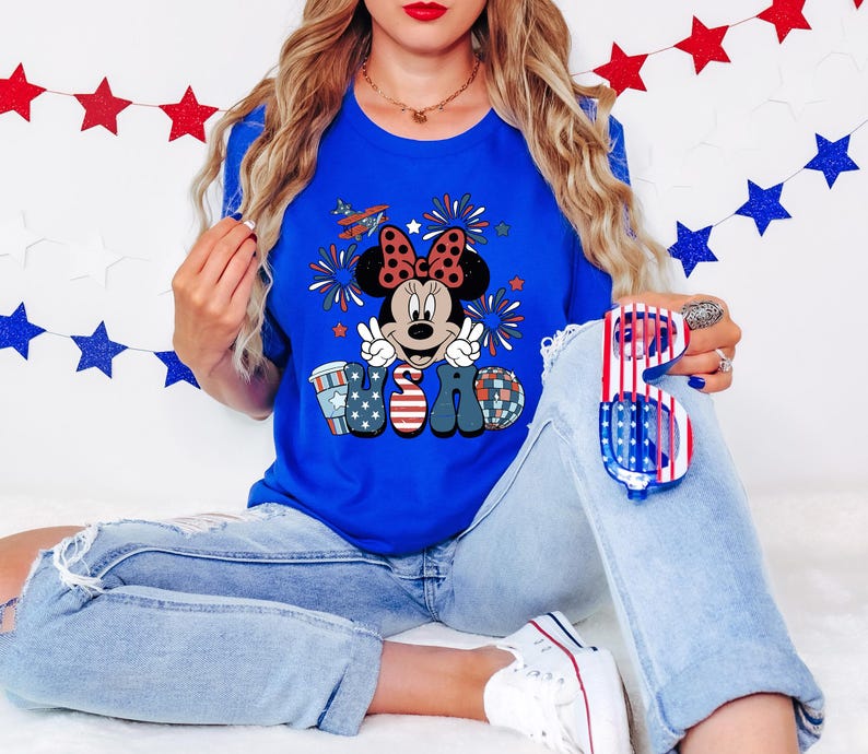 May include: A royal blue t-shirt featuring a Minnie Mouse graphic with a red bow, peace signs, and the letters "USA" in patriotic colors. The person is wearing light blue jeans and holding red, white, and blue sunglasses.