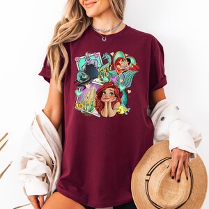 A woman wearing a burgundy t-shirt with a colorful graphic of Disney characters, including mermaids, is holding a straw hat and standing against a white background.