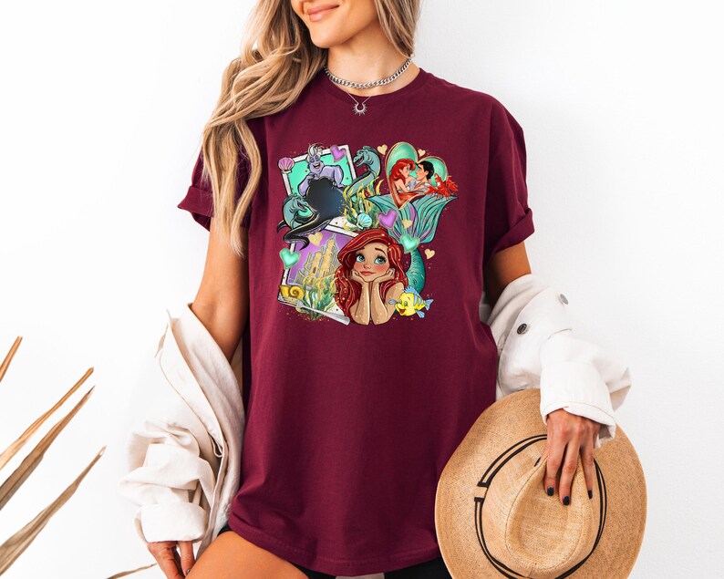 A woman wearing a burgundy t-shirt with a colorful graphic of Disney characters, including mermaids, is holding a straw hat and standing against a white background.