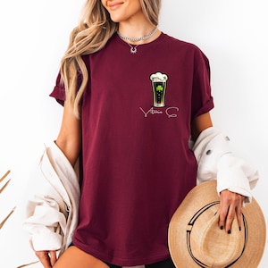 A woman wearing a burgundy t-shirt with a graphic of a beer glass and the text "Amana" printed on it, holding a straw hat in her hand.