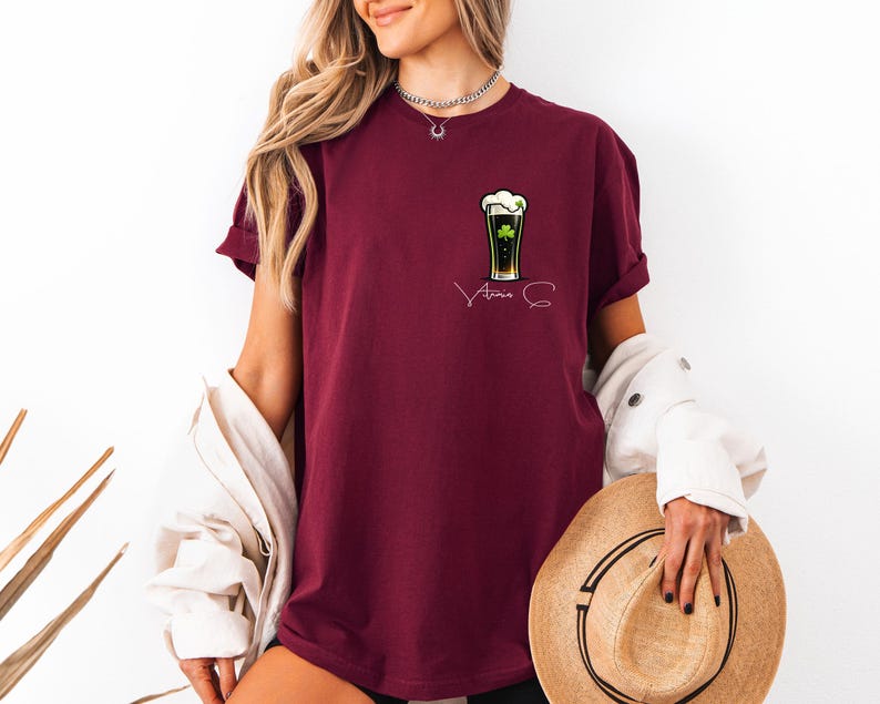 A woman wearing a burgundy t-shirt with a graphic of a beer glass and the text "Amana" printed on it, holding a straw hat in her hand.
