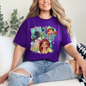 A woman wearing a purple t-shirt with a colorful graphic of Disney characters is sitting on a couch.