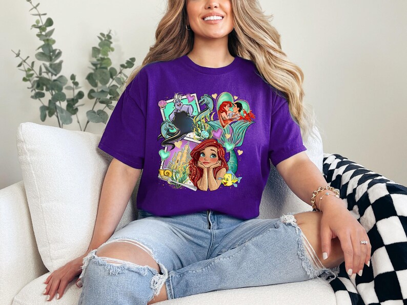 A woman wearing a purple t-shirt with a colorful graphic of Disney characters is sitting on a couch.