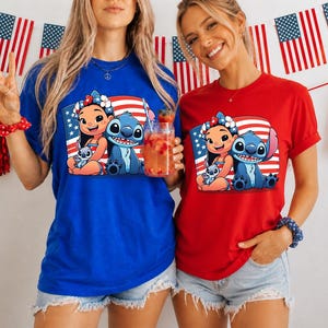 May include: Two people wearing blue and red t-shirts featuring a cartoon image of Lilo and Stitch with an American flag background. The shirts are paired with denim shorts. The background includes American flags and festive decorations.
