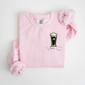 A pink sweatshirt with a graphic of a beer mug and a clover leaf on the front, along with the text "Atria" printed on it.