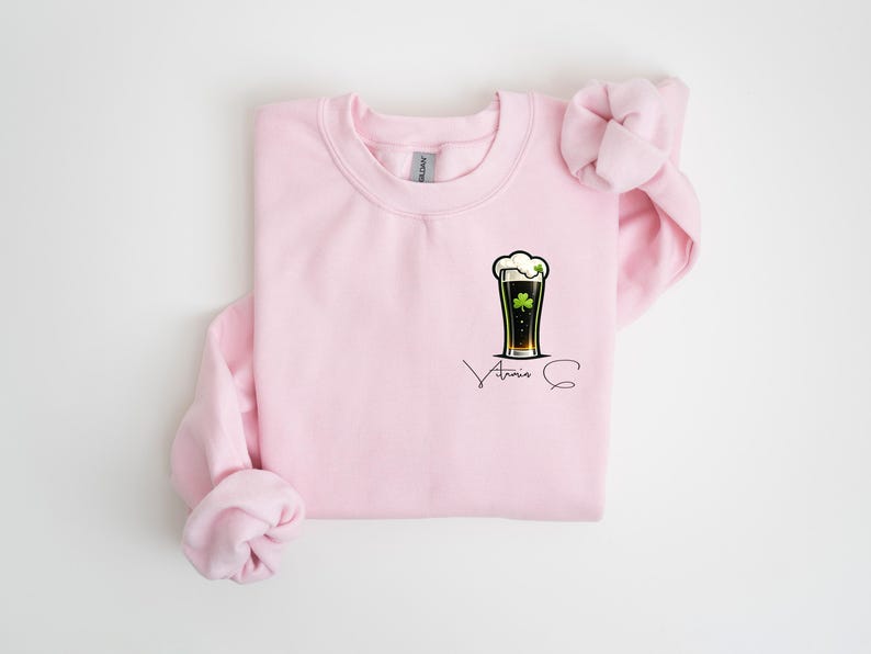 A pink sweatshirt with a graphic of a beer mug and a clover leaf on the front, along with the text "Atria" printed on it.