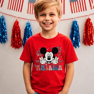 May include: A red t-shirt featuring a graphic of Mickey Mouse with peace signs and the word "AMERICA" in patriotic colors. The shirt is worn by a smiling child, with American flags and red and blue decorations in the background.