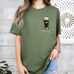 A woman wearing a green t-shirt with a graphic of a beer and a clover, standing in front of a wooden wall with a circular design.