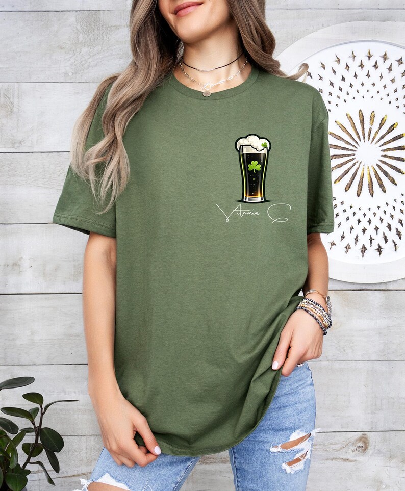 A woman wearing a green t-shirt with a graphic of a beer and a clover, standing in front of a wooden wall with a circular design.