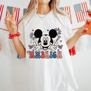 May include: White t-shirt featuring a Mickey Mouse graphic with the word "AMERICA" in red, white, and blue. The design includes fireworks and stars. The shirt is part of a patriotic theme, perfect for Independence Day celebrations.