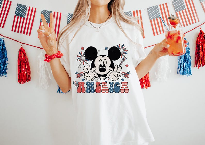 May include: White t-shirt featuring a Mickey Mouse graphic with the word "AMERICA" in red, white, and blue. The design includes fireworks and stars. The shirt is part of a patriotic theme, perfect for Independence Day celebrations.