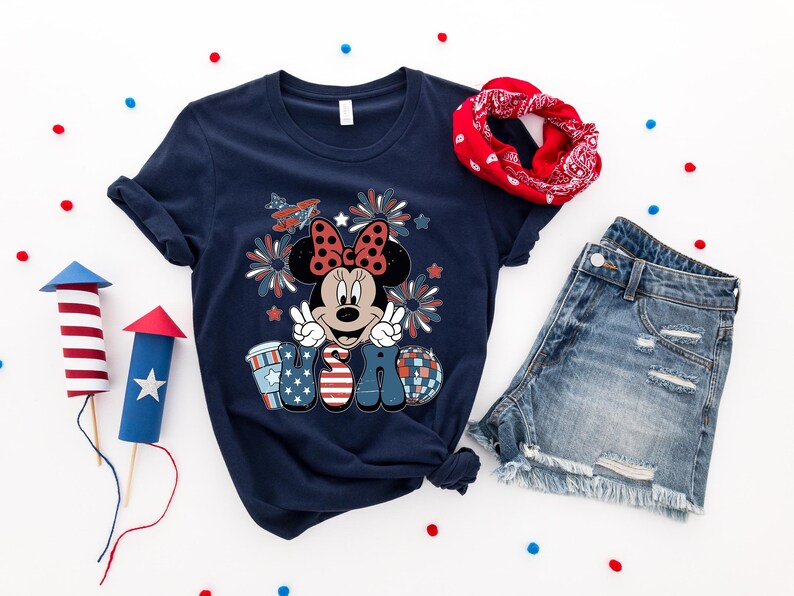 May include: Navy blue t-shirt featuring Minnie Mouse with a patriotic design, peace sign, and the word "USA". Includes denim shorts and a red bandana. Decorative red, white, and blue confetti and paper rockets complete the festive ensemble.