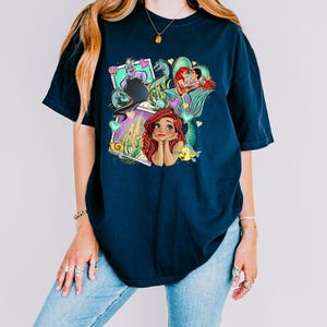 A woman wearing a blue t-shirt with a colorful graphic design featuring characters from the Disney animated film "The Little Mermaid".