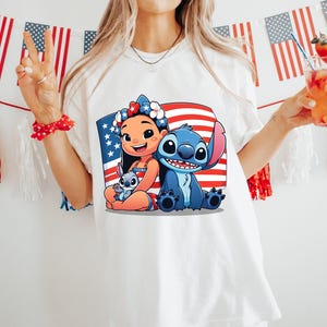 May include: White t-shirt featuring a graphic of Lilo and Stitch with an American flag background. The design includes Lilo, Stitch, and a small Stitch. The shirt is worn by a person holding a drink with a strawberry garnish.