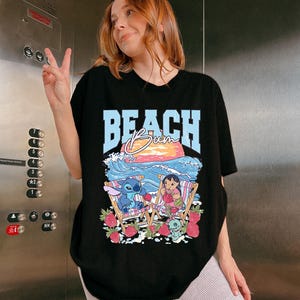 A woman wearing a black t-shirt with a graphic design of a cartoon character and the word "BEACH" printed on it, smiling and making a peace sign gesture.