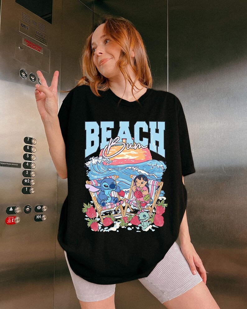 A woman wearing a black t-shirt with a graphic design of a cartoon character and the word "BEACH" printed on it, smiling and making a peace sign gesture.