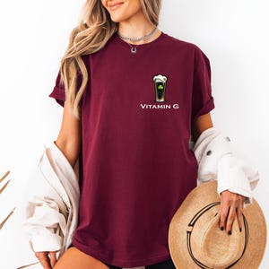 A woman wearing a burgundy t-shirt with a graphic of a vitamin G bottle and holding a straw hat.