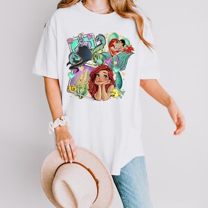 A woman wearing a white t-shirt with a colorful graphic of mermaid characters and holding a beige hat.