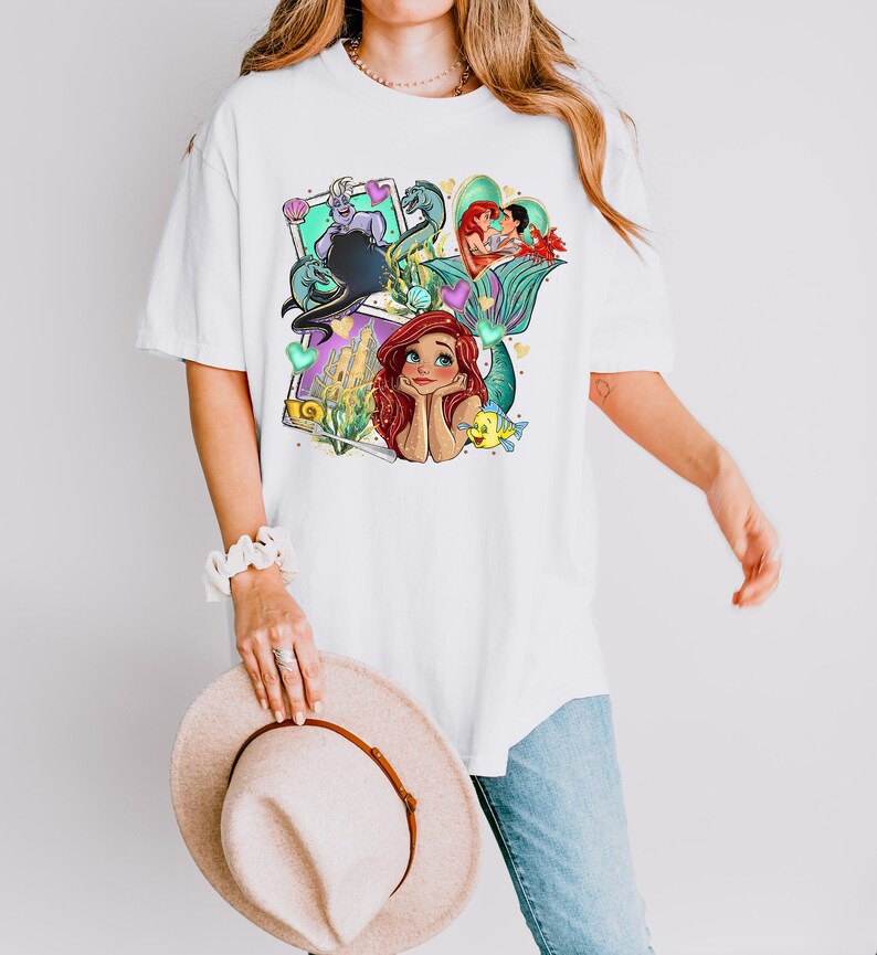 A woman wearing a white t-shirt with a colorful graphic of mermaid characters and holding a beige hat.