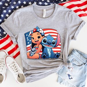 May include: A gray t-shirt featuring a colorful graphic of Lilo and Stitch with an American flag background. The shirt is paired with denim shorts and white sneakers, with an American flag in the background.