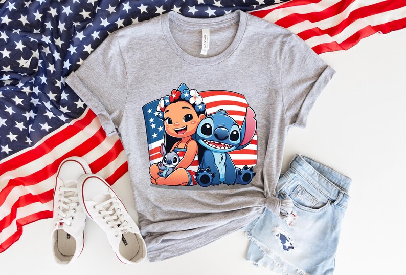 May include: A gray t-shirt featuring a colorful graphic of Lilo and Stitch with an American flag background. The shirt is paired with denim shorts and white sneakers, with an American flag in the background.