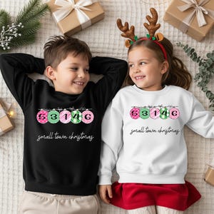 May include: Two children wearing matching sweatshirts with Christmas ornament designs. The ornaments are pink and green with the numbers 53146. The text "small town christmas" is below the ornaments. One sweatshirt is black, the other is white.