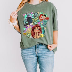 A woman wearing a green t-shirt with a colorful graphic of mermaid characters and a castle.