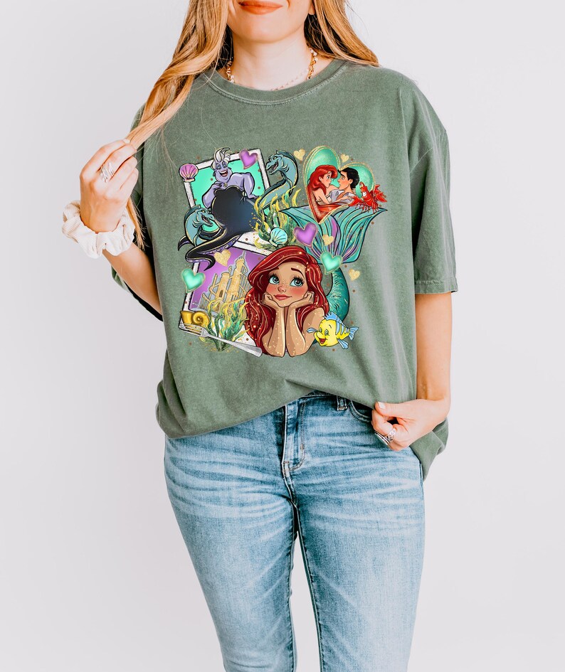 A woman wearing a green t-shirt with a colorful graphic of mermaid characters and a castle.
