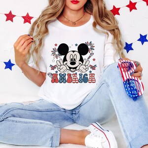 May include: White t-shirt featuring a graphic of Mickey Mouse with peace signs, fireworks, and the word "AMERICA" in red, white, and blue. The design is patriotic, with stars and stripes elements. The shirt is paired with blue jeans.