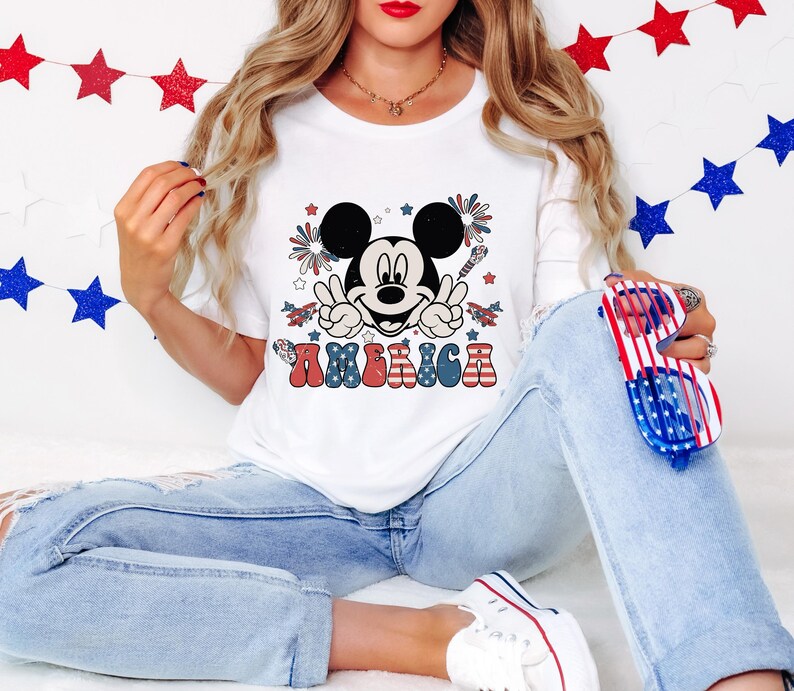 May include: White t-shirt featuring a graphic of Mickey Mouse with peace signs, fireworks, and the word "AMERICA" in red, white, and blue. The design is patriotic, with stars and stripes elements. The shirt is paired with blue jeans.