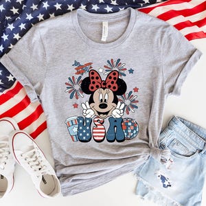 May include: A light gray t-shirt featuring a Minnie Mouse graphic with a red bow, peace signs, and the word "USA" in patriotic colors. The shirt is paired with denim shorts and white sneakers, with an American flag in the background.